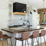 Modern kitchen with wooden island and stools, featuring a black range hood and marble backsplash.