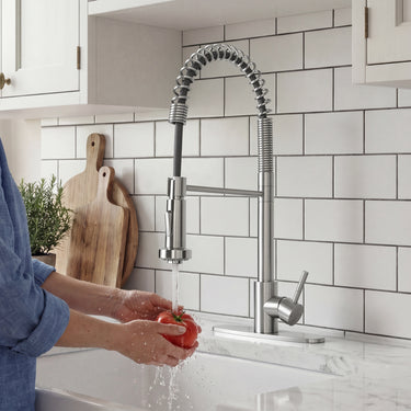 Person washing a tomato under a modern kitchen faucet with a tiled backsplash.