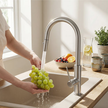 Person washing grapes under a kitchen faucet with a bowl of fruit on the counter.