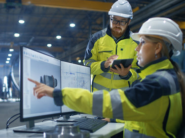 Two engineers in a factory setting, one pointing at a computer screen and the other holding a tablet.